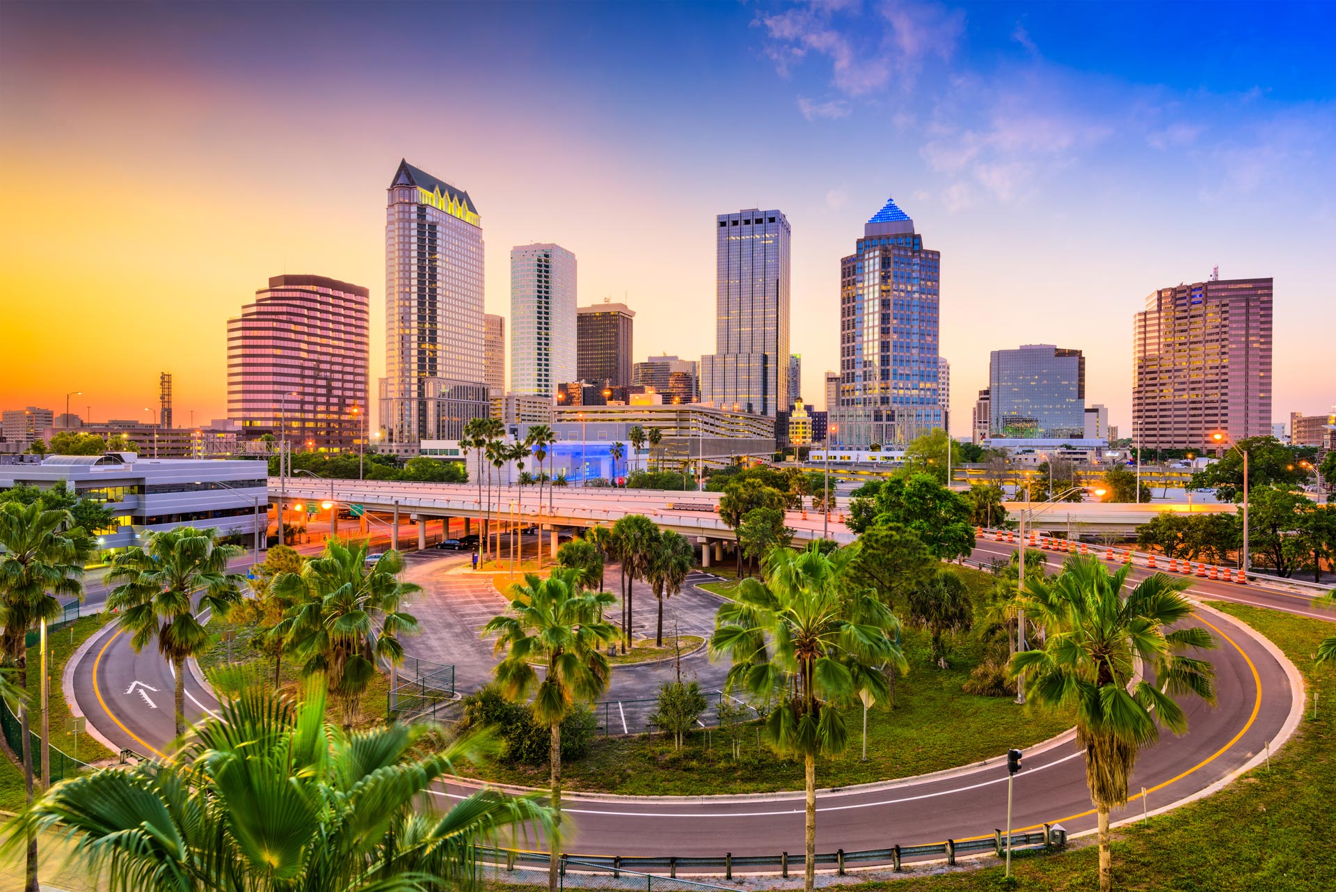The image shows a vibrant city skyline at sunset with a large, colorful neon sign, palm trees, and a clear sky.