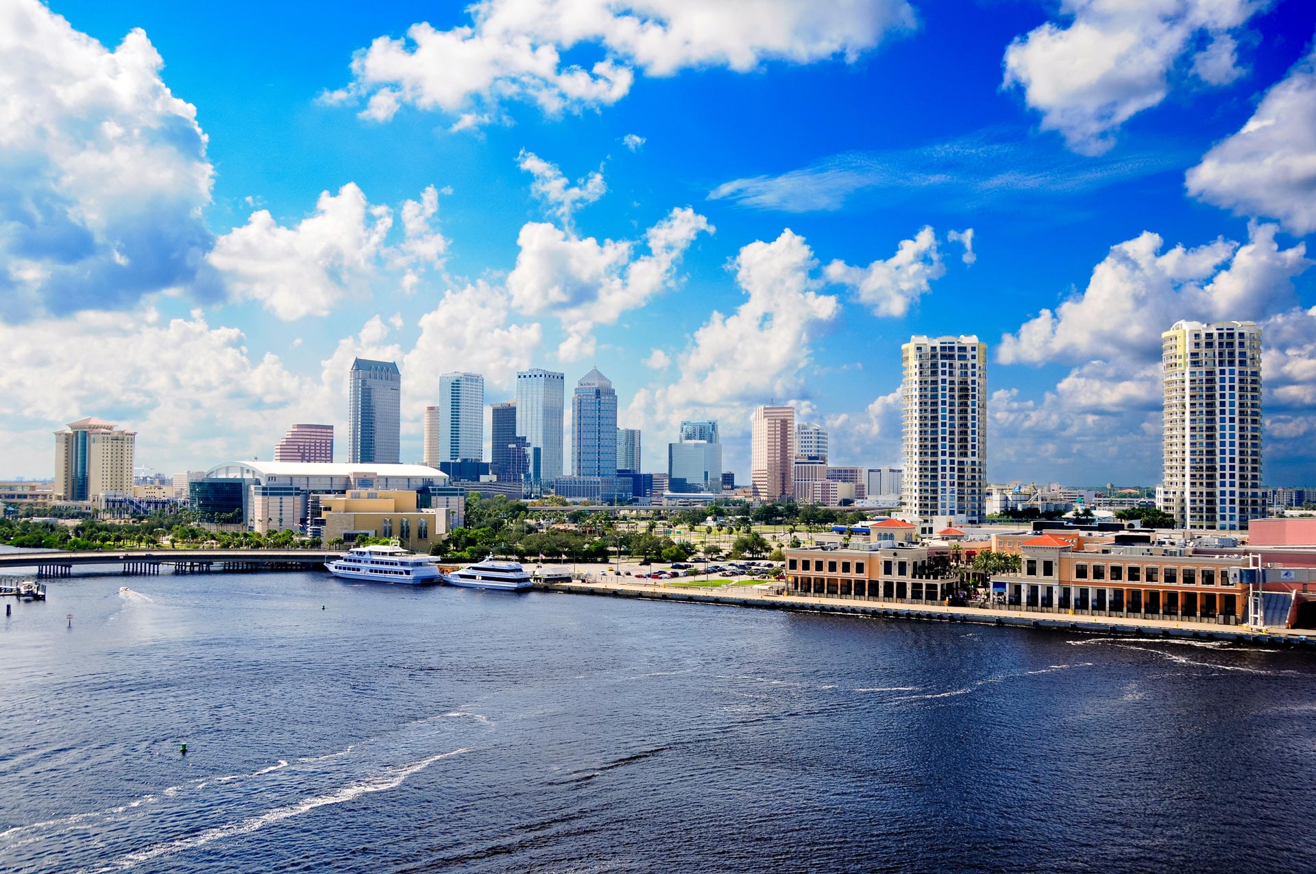 The image shows a panoramic view of a city skyline with a river, boats, buildings, and a clear blue sky with some clouds.